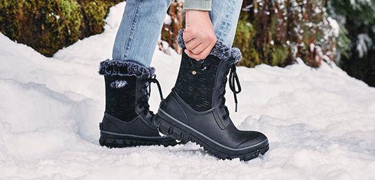 Black Bogs winter boots with fur lining and rugged soles worn in snowy forest terrain; person adjusting laces while wearing jeans and a green sweater, showcasing insulated waterproof footwear for cold weather.