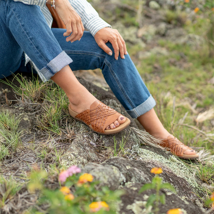 Person sitting on rocky grass wearing brown woven Taos Guru sandals with rolled‑up jeans, shown in a spring outdoor setting with wildflowers.
