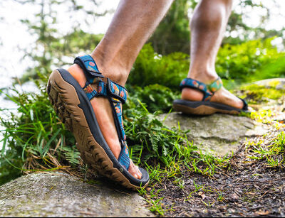 Man hiking on a rocky, grassy trail wearing men’s Teva hiking sandals with colorful straps and rugged traction soles in a forest setting.