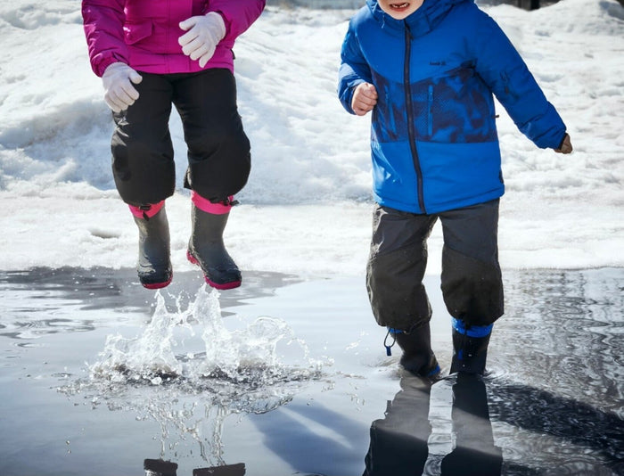 Two children playing in snowy puddles wearing waterproof Kamik winter boots; pink-trimmed and black styles shown in active outdoor use, highlighting traction, insulation, and durability for cold weather play.
