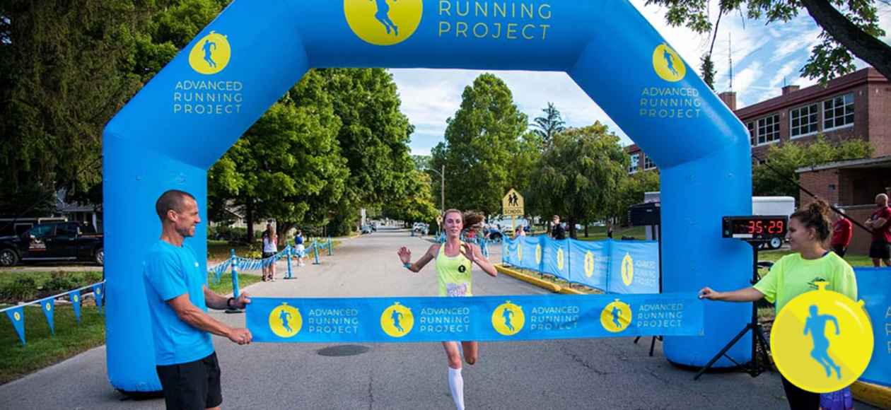 “Runner breaking the finish-line ribbon under a blue ‘Advanced Running Project’ arch during the Virtual Race Series 2020