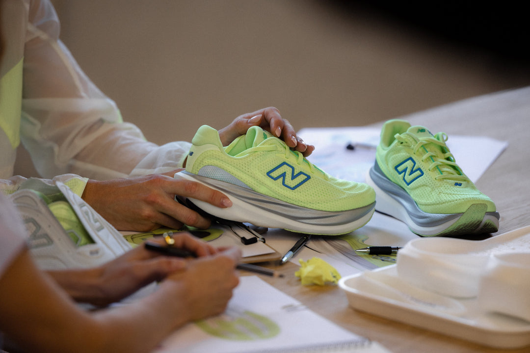 Person examining a neon green New Balance 1080v15 running shoe in a design studio with sketches and tools on the table