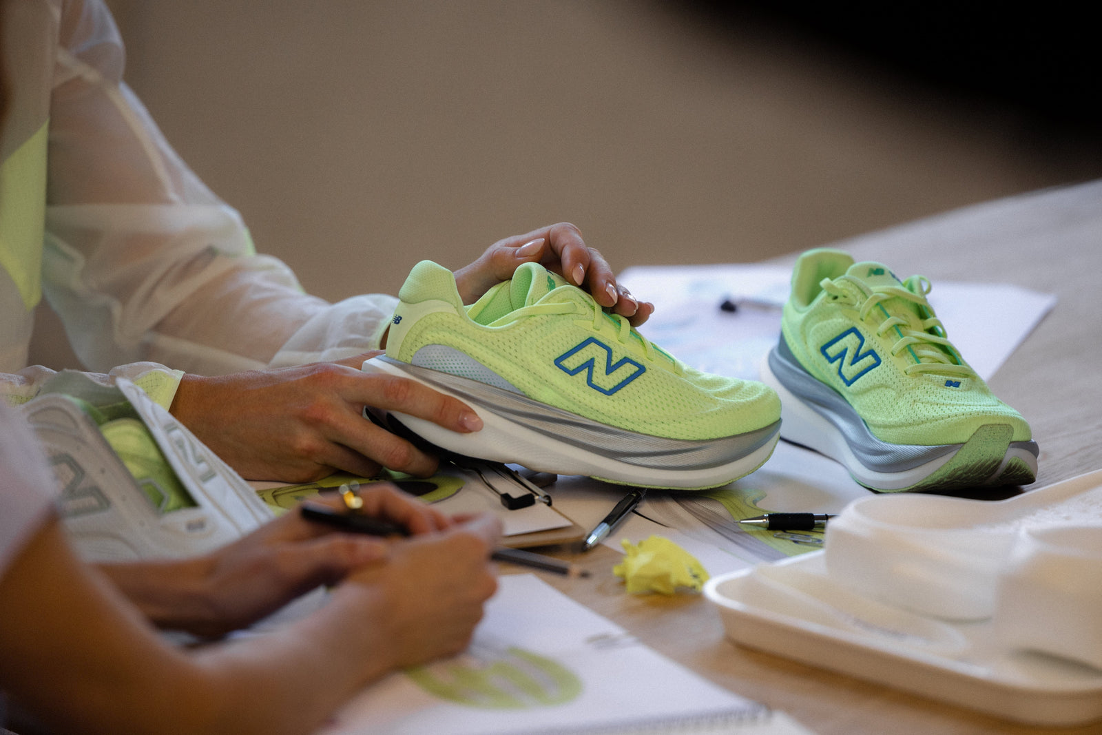 Person examining a neon green New Balance 1080v15 running shoe in a design studio with sketches and tools on the table