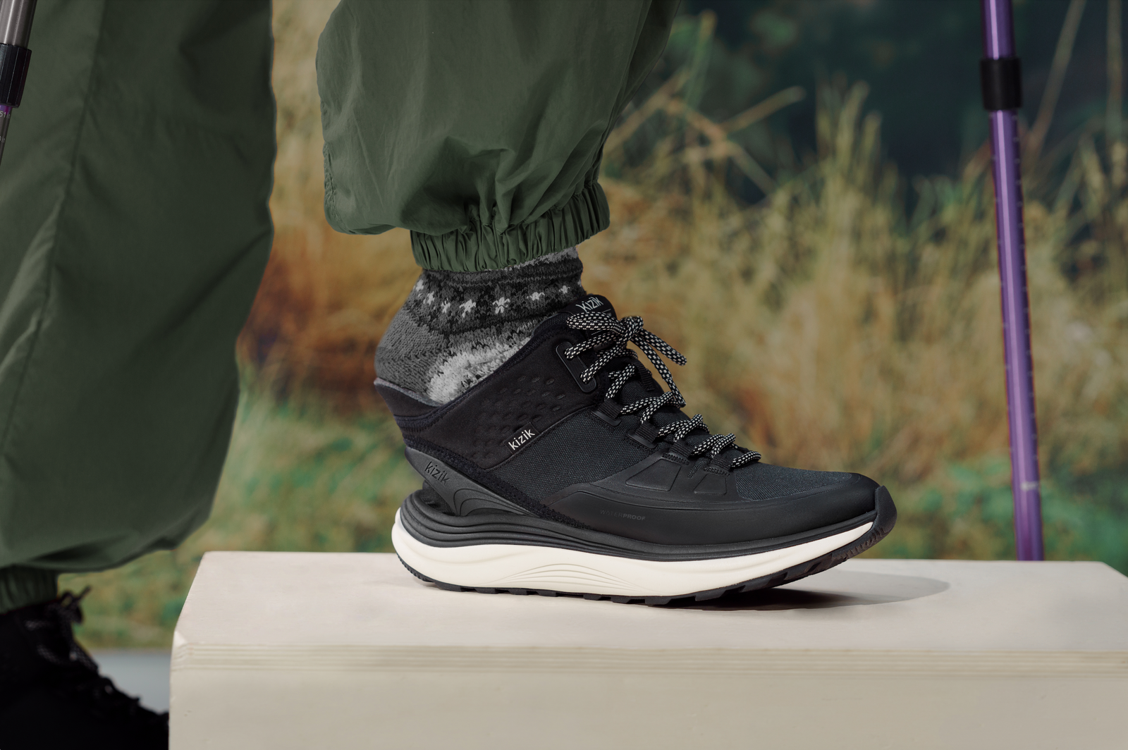 Woman wearing Kizik London AT Mid WP hiking boots in Black/Whitecap Gray with snowflake socks and green pants; close-up shows hands-free step-in design in an outdoor setting.
