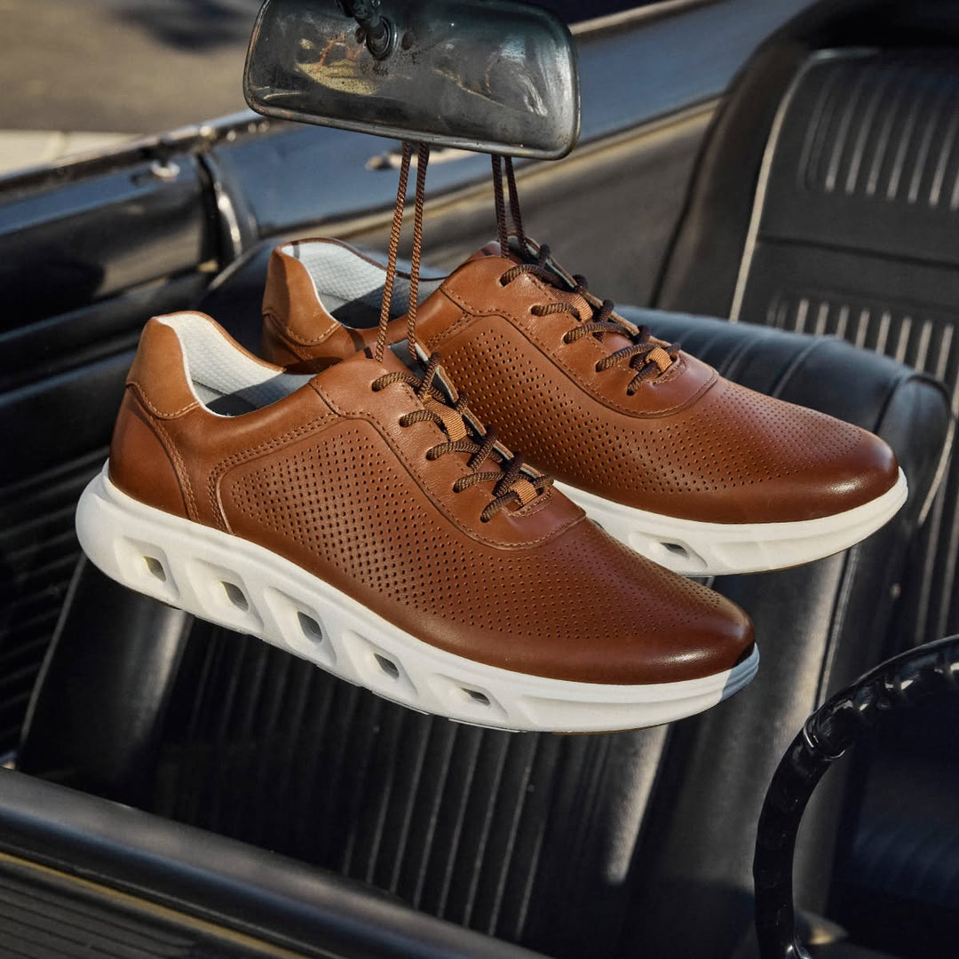 Tan perforated leather Johnston & Murphy Reactivate U-Throat sneakers with white cushioned soles, hanging by their laces from the rearview mirror of a vintage car