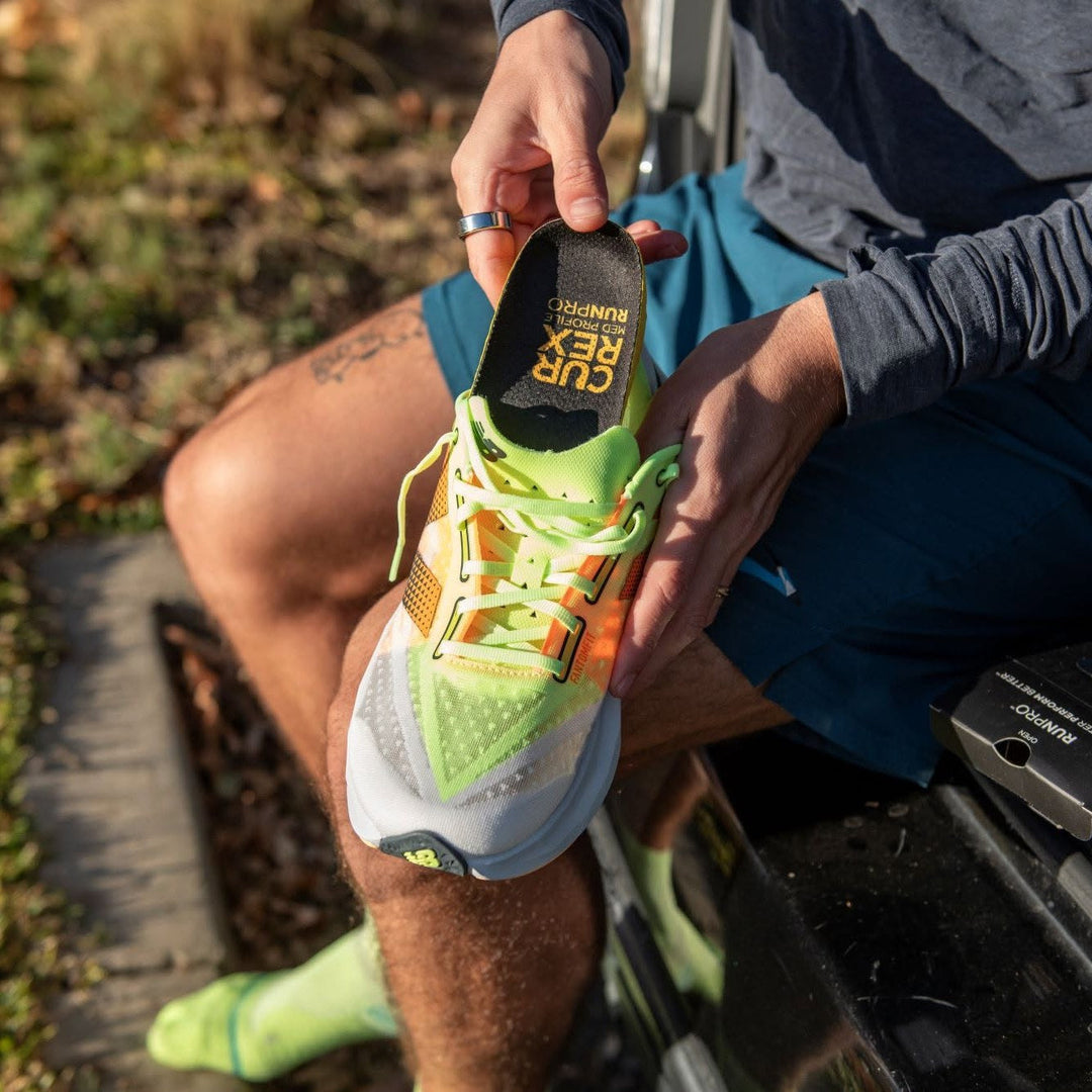 Runner in neon green socks inserting a CURREX RunPro Med Profile insole into a matching green and orange running shoe outdoors