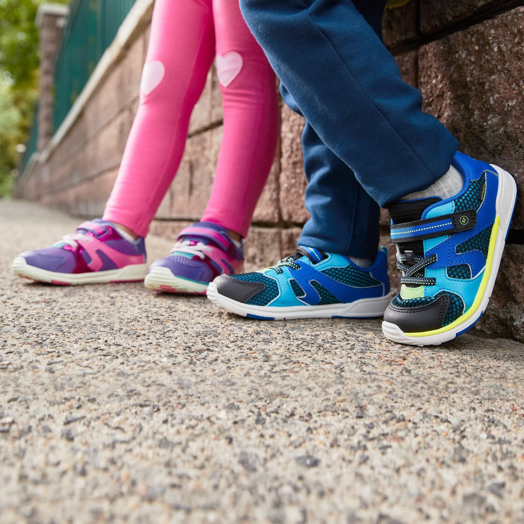 Two toddlers in blue and purple Stride Rite Velocity sneakers leaning against a brick wall