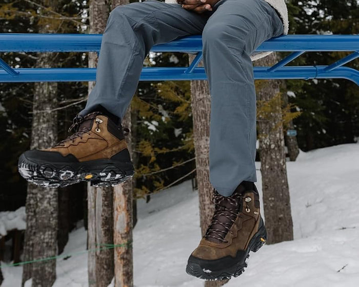 Man wearing brown Merrell hiking boots with black soles and visible snow, sitting on blue metal structure in snowy forest; boots designed for winter traction, insulation, and outdoor durability.