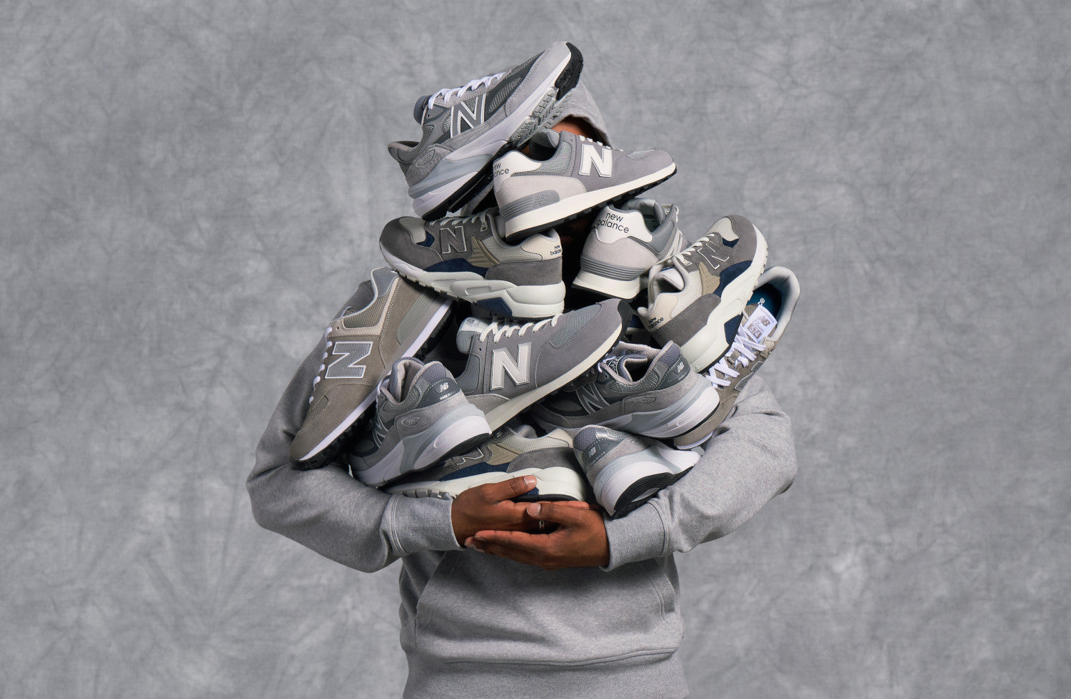 Person holding stacked gray New Balance sneakers, wearing light gray sweatshirt against textured gray background