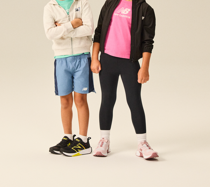 Two kids wearing New Balance athletic apparel, featuring black New Balance kids’ sneakers with yellow accents and white‑and‑pink New Balance running shoes.