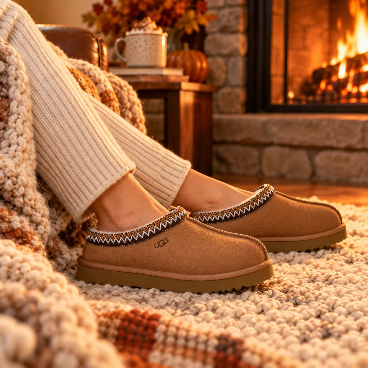 Woman relaxing by a fireplace in chestnut UGG Tasman II slippers with a cozy knit blanket and hot cocoa